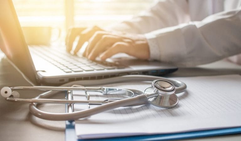 Stethoscope placed on a notebook with a person typing on a laptop in the background.